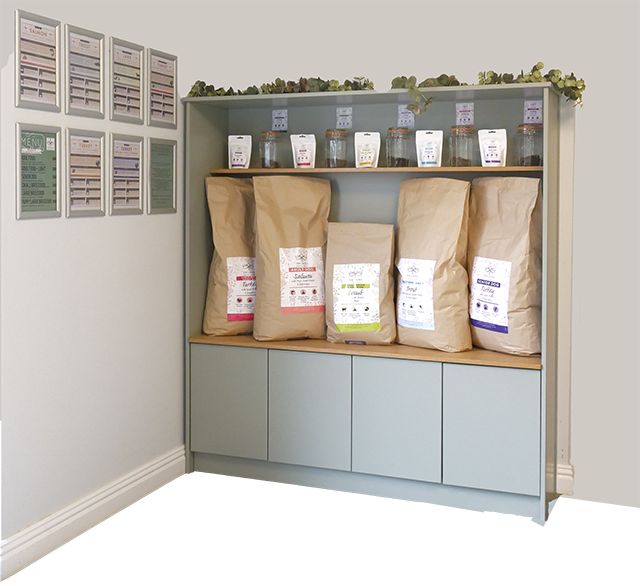 Display dresser in reception with a top shelf containing the five treat typesin bags with glass jars for sampling. Five large food bags are displayed underneath. On the wall there are satin silver frames with more information